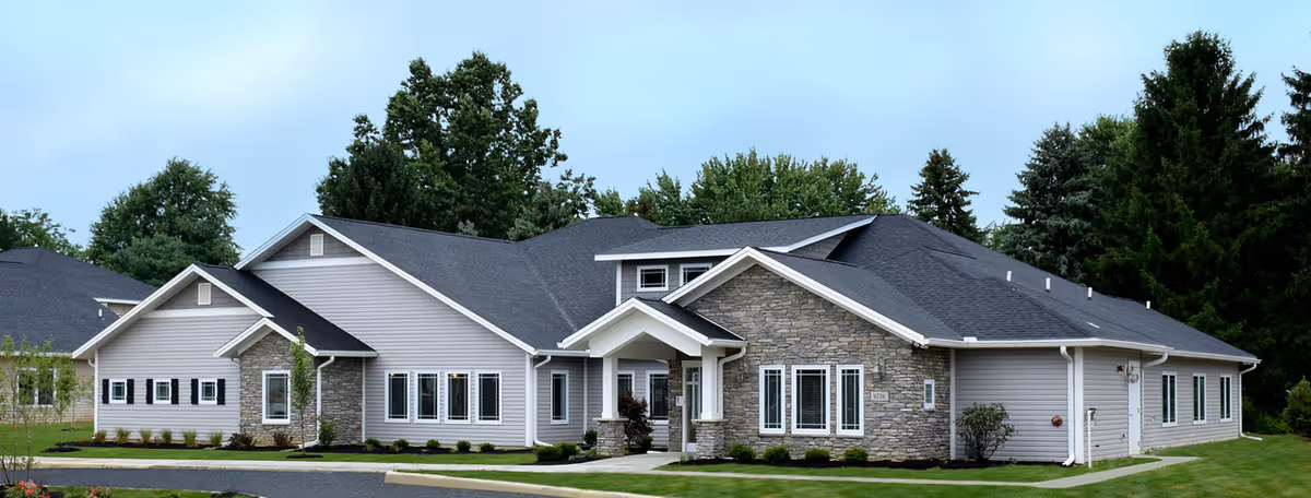 Single-story stone and gray-siding senior living building with a front entrance, lawn, and surrounding trees.