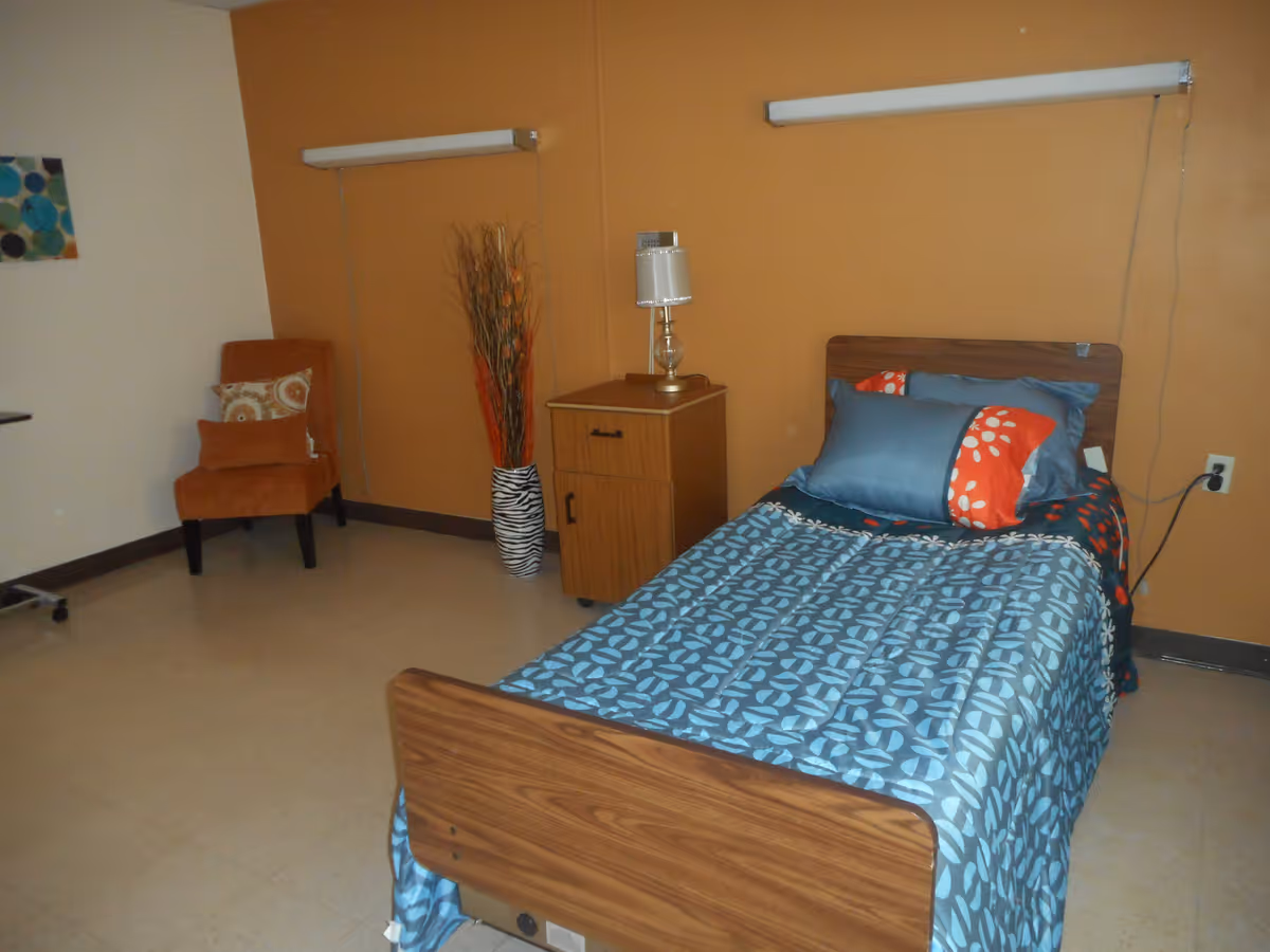 A simple bedroom with a single bed covered in blue and orange patterned bedding, a wooden nightstand with a lamp, an orange upholstered chair with a decorative pillow, and a tall vase with dried branches against an orange accent wall.