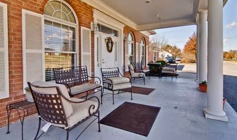 Covered outdoor porch area with cushioned metal chairs and benches arranged along a brick wall with white window shutters and a white door decorated with a wreath. The porch has large white columns and overlooks a driveway and some trees in the background.