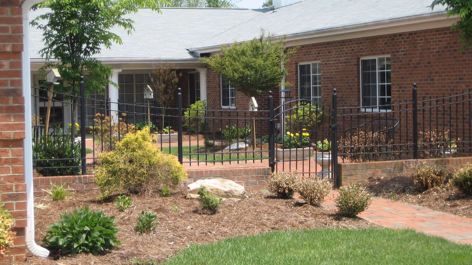 Outdoor view of a brick building with a black metal fence and gate surrounding a landscaped garden area with bushes, small trees, and a brick pathway leading to the entrance.