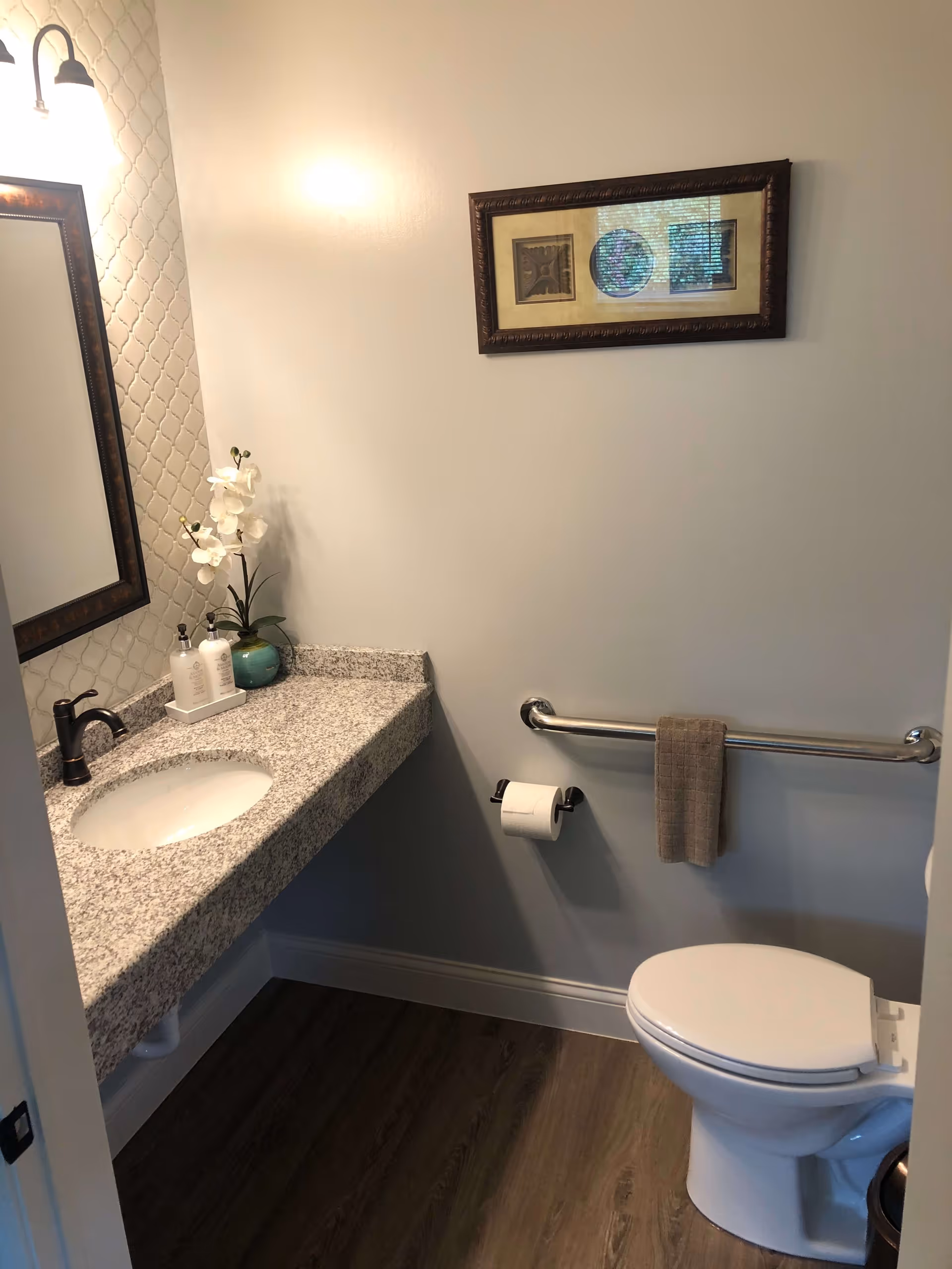 A small bathroom with a white toilet, a granite countertop with an under-mount sink, a bronze faucet, a mirror with a dark wooden frame, a wall-mounted light fixture, a decorative framed picture on the wall, a hand towel hanging on a metal grab bar, a toilet paper holder, and a small green vase with white flowers on the countertop.
