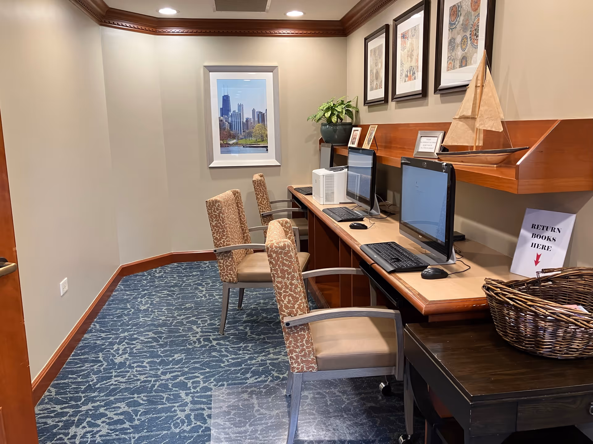 A small computer room with two desktop computers on a wooden desk, two patterned chairs, a basket with books, a sign that says 'RETURN BOOKS HERE', framed artwork on the wall, and a model sailboat on a shelf.