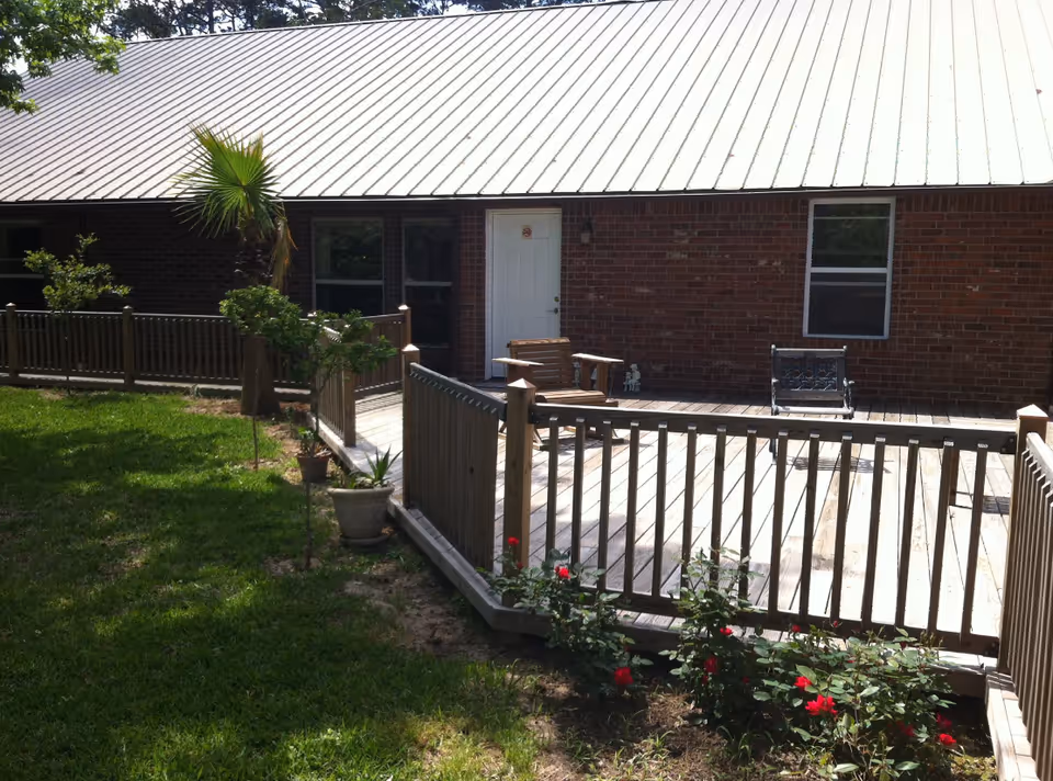 Outdoor view of a senior care facility showing a wooden deck with railing, a wooden chair, a metal bench, and a brick building with a white door and windows. There are small plants and flowers along the edge of the deck and a grassy lawn in the foreground.