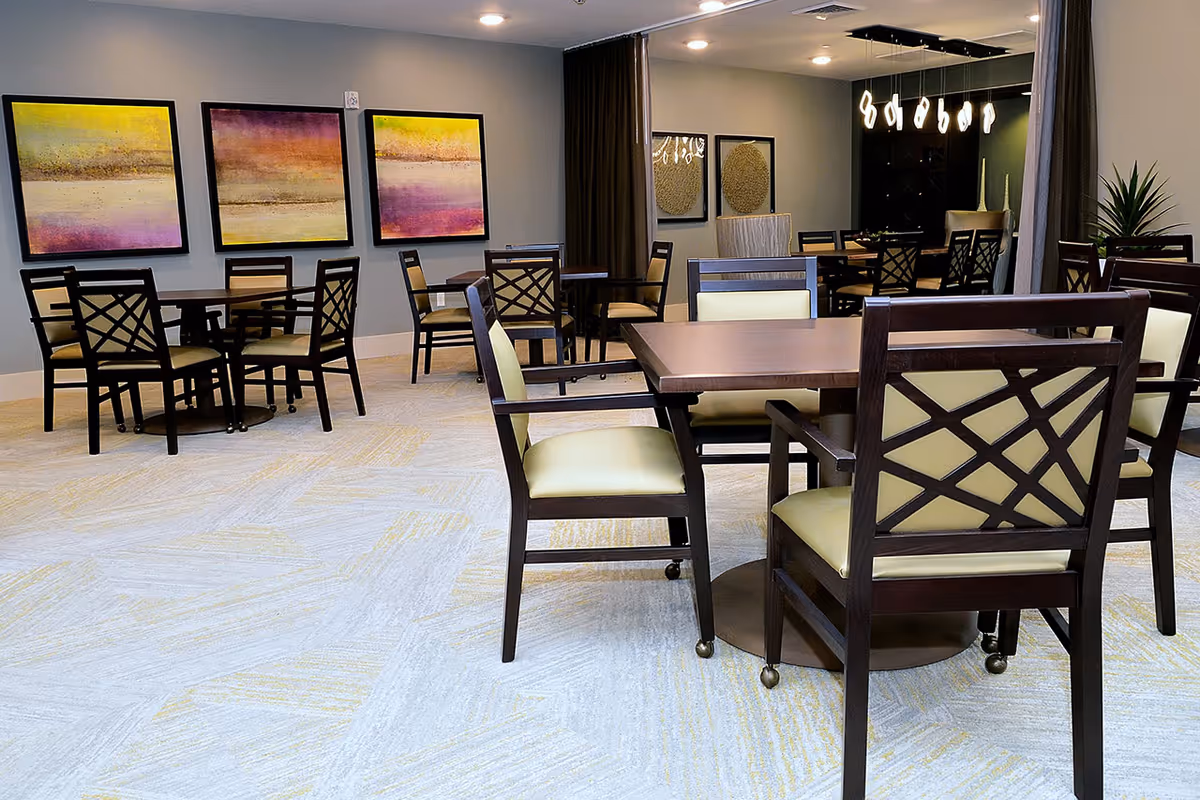 A modern dining room with several wooden tables and chairs featuring light yellow cushions. The room has light-colored carpet flooring with subtle yellow patterns, gray walls adorned with three colorful abstract paintings, and a large mirror reflecting part of the room. A contemporary chandelier with multiple pendant lights hangs in the background, and a potted plant is visible on the right side.