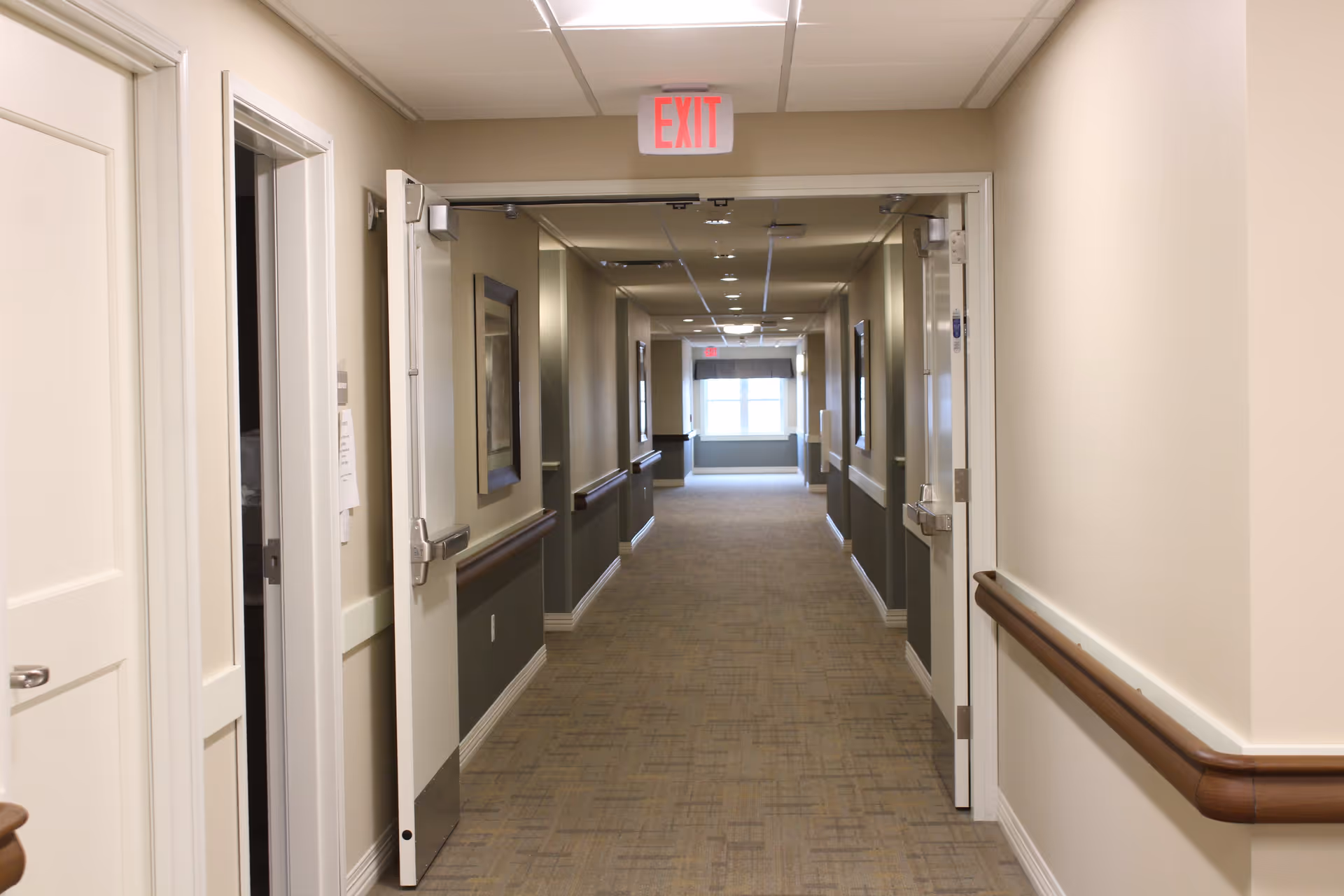 A long, well-lit hallway in a senior living facility with beige walls, carpeted floor, handrails on both sides, and double doors open in the foreground. There are framed pictures on the walls and an exit sign above the doors. A window at the end of the hallway lets in natural light.