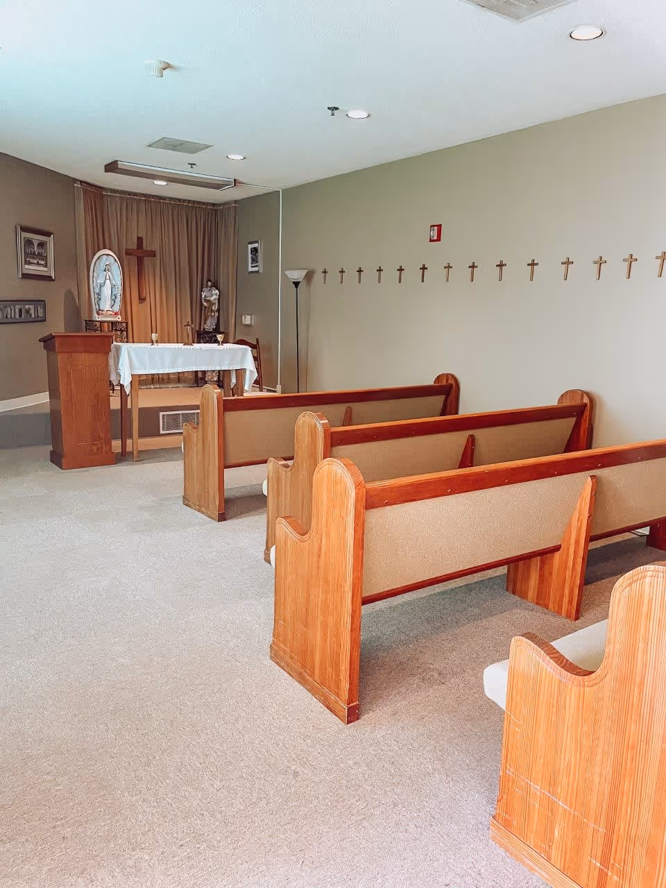 Interior of a small chapel with wooden pews arranged in rows facing an altar. The altar is covered with a white cloth and has religious statues and a wooden cross behind it. The walls are decorated with multiple small crosses and framed pictures. The room has beige carpeting and neutral-colored walls.