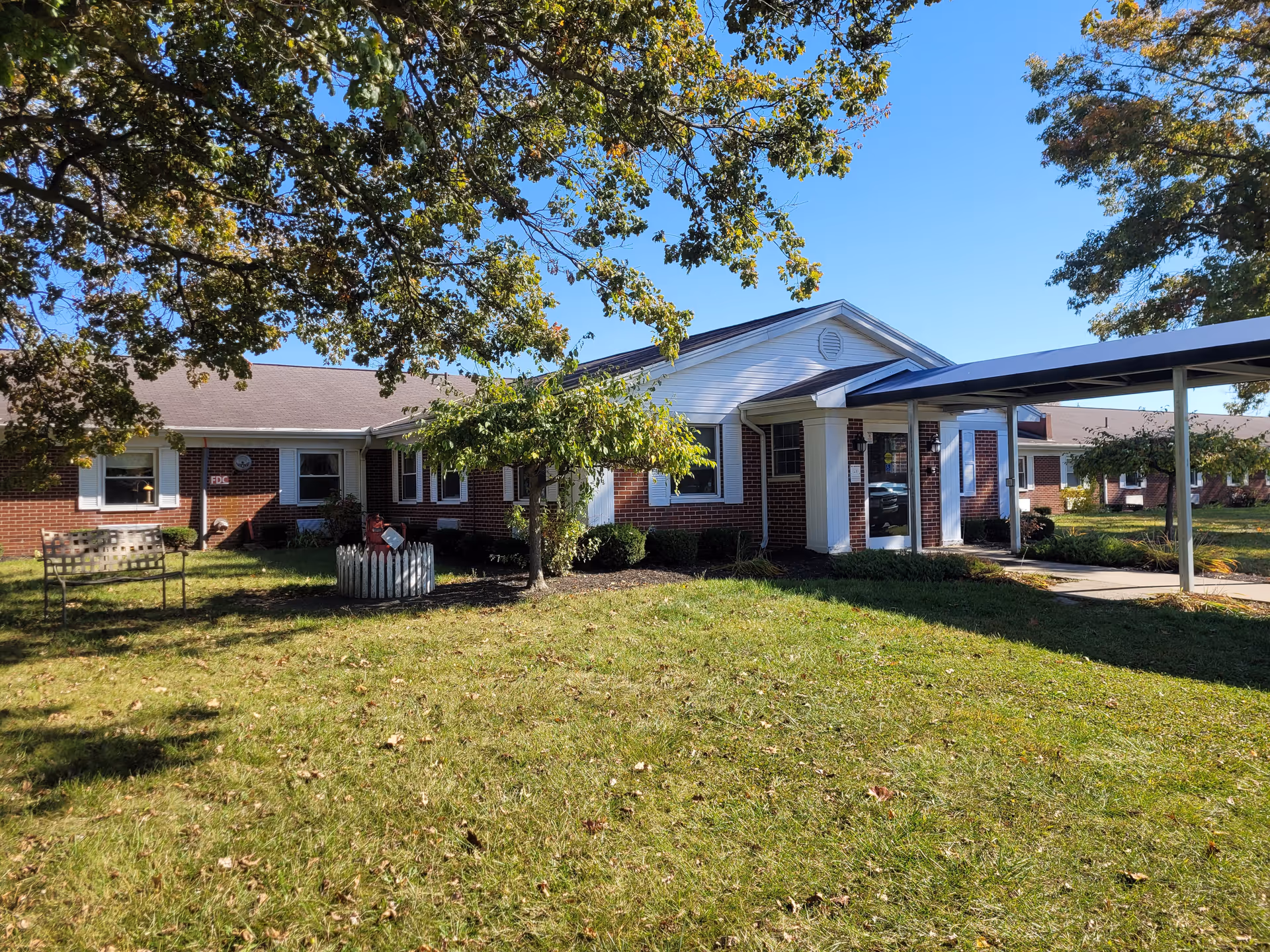 Exterior view of a single-story brick building with white trim, surrounded by green grass and trees under a clear blue sky. There is a covered walkway leading to the entrance, and a bench and small fenced garden area are visible on the lawn.