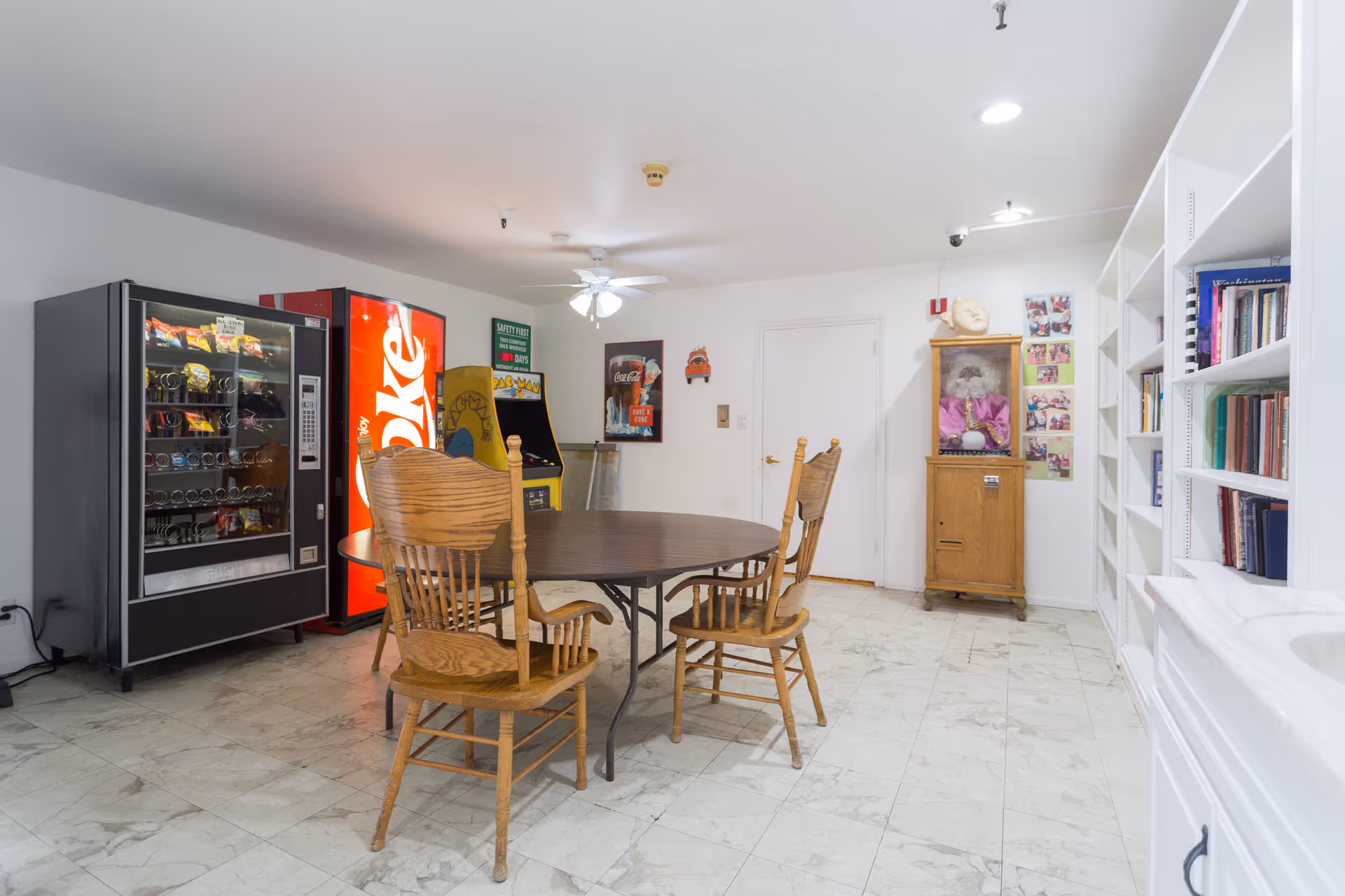 A common room with a round wooden table and four wooden chairs. Against the wall are a snack vending machine, a Coca-Cola vending machine, and an arcade game. There is a ceiling fan with lights, a wooden cabinet with a doll inside, and bookshelves filled with books on the right side.