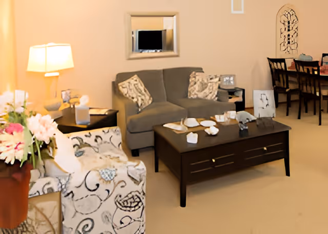 Living area with a loveseat, patterned armchair, central coffee table, lamp, and a dining table visible in the background.