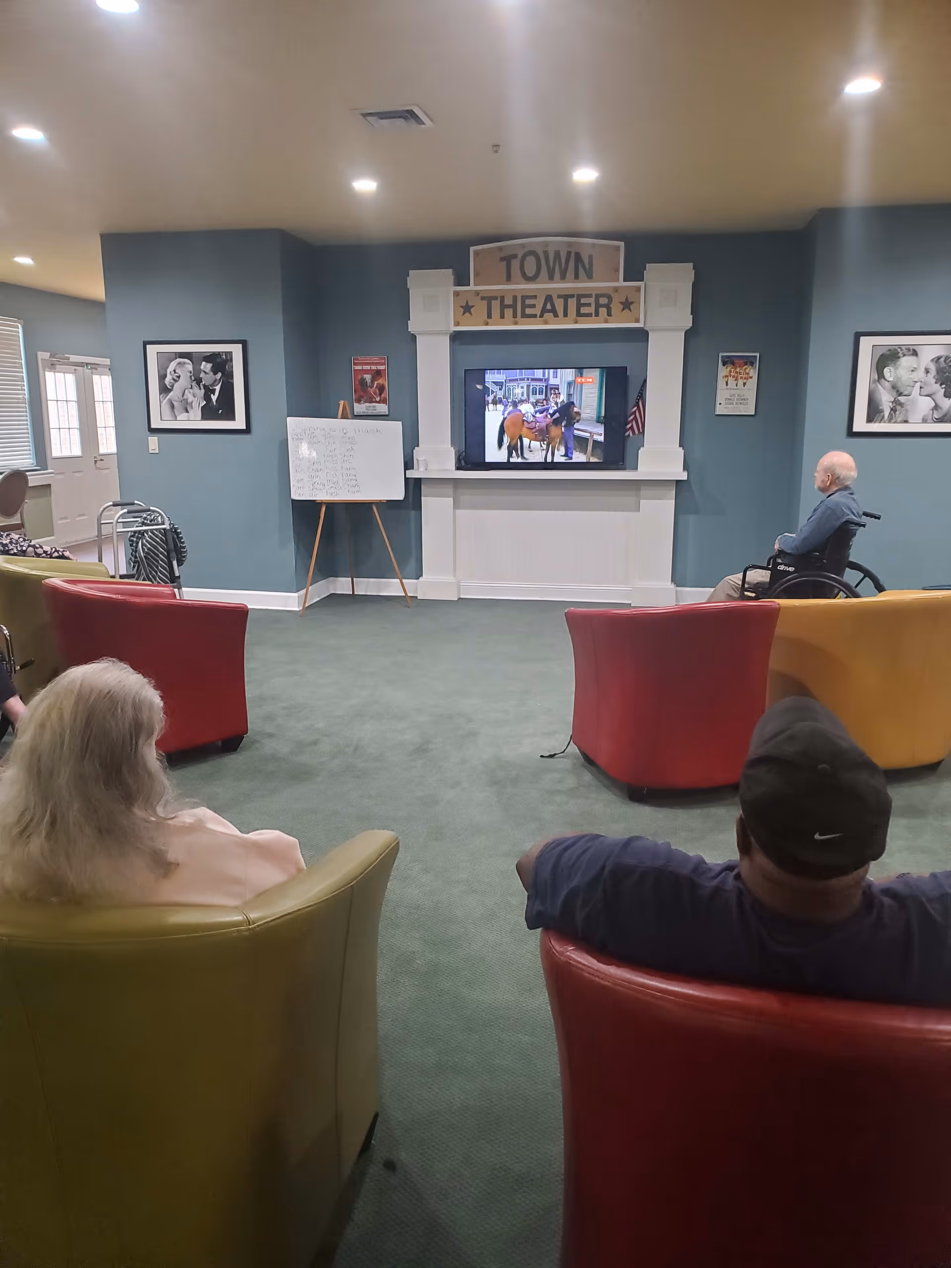 A group of elderly people seated in colorful armchairs and a wheelchair watching a television in a room labeled 'Town Theater'. The room has blue walls, framed black and white photos, a whiteboard with writing, and a green carpet.