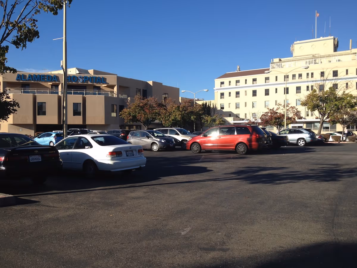 Parking lot with several cars parked in front of a large hospital building under a clear blue sky. The building has a sign that reads 'ALAMEDA HOSPITAL'. Trees with autumn foliage are visible near the building.