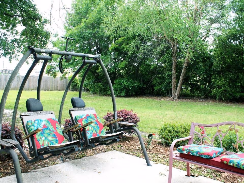 Backyard patio with two cushioned swing seats and a cushioned metal bench overlooking a grassy yard and trees.
