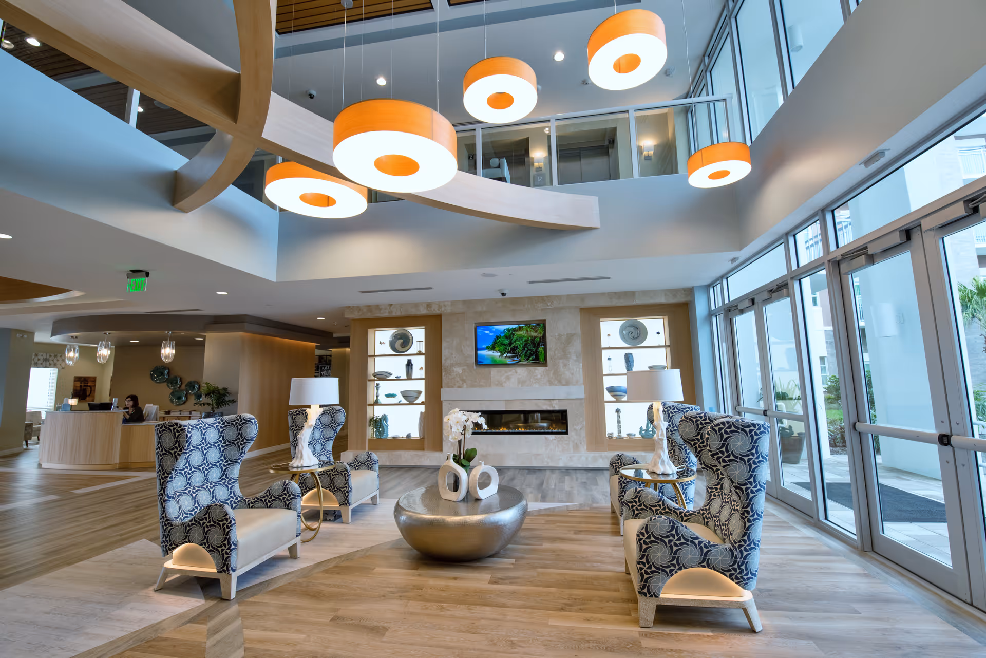 A modern and spacious senior living facility lobby with high ceilings and large windows allowing natural light. The seating area includes four patterned armchairs arranged around a round metallic coffee table with decorative vases. Two side tables with lamps flank the chairs. The back wall features a built-in fireplace with a mounted TV above it and decorative shelves on either side. The reception desk is visible in the background to the left, and large glass doors lead outside on the right.