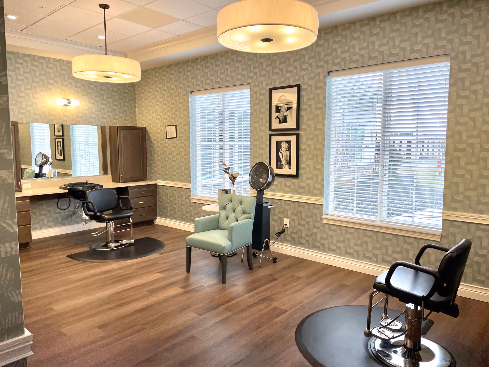 Interior of a hair salon area in a senior living facility with two black salon chairs on black mats, a teal armchair, a hair dryer on a stand, two large windows with blinds, patterned wallpaper, framed black and white photos on the wall, and two large ceiling lights.