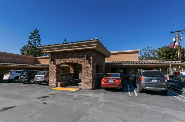 Exterior view of a single-story building with a stone entrance canopy and several parked cars in front under a clear blue sky.