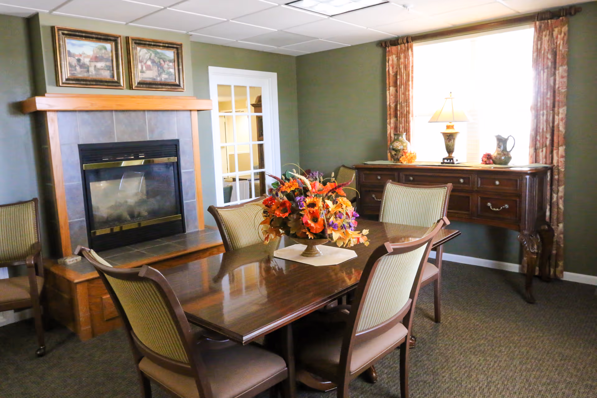 A cozy dining room with a wooden dining table surrounded by six chairs. A floral centerpiece sits on the table. In the background, there is a fireplace with a wooden mantel and two framed paintings above it. To the right, a wooden sideboard with decorative items and a lamp is placed in front of a window with floral curtains.