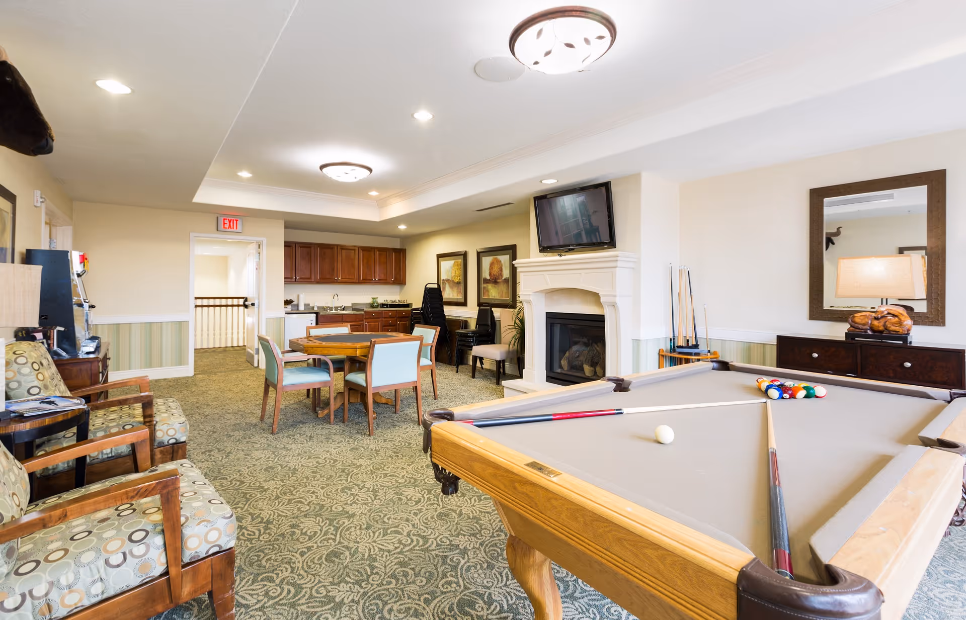 A senior living facility game room featuring a pool table with cues and balls, a round table with chairs, a fireplace with a mounted TV above it, a small kitchenette with cabinets and a sink, and several upholstered chairs along the wall. The room has patterned carpet, recessed lighting, and a large mirror above a dark wooden cabinet.
