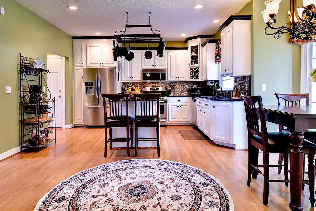 A bright kitchen with white cabinets, stainless steel refrigerator and oven, a hanging pot rack with pots and pans, a wooden dining table with chairs, and a round patterned rug on a wooden floor. The walls are painted green and there is a window with a floral valance.