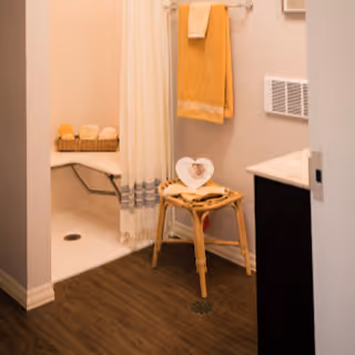 A bathroom with a walk-in shower featuring a built-in bench and a white curtain with gray trim. A wicker basket with towels is placed on the bench. A wooden stool with a heart-shaped photo frame sits on the wood-look floor. A yellow towel hangs on a wall-mounted towel rack next to a vent and a countertop.