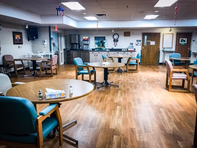 A spacious dining room with several round tables and chairs arranged around the room. The floor is wood, and the walls have a few framed pictures and a clock. There is a counter area with appliances and cabinets in the background, and the ceiling has fluorescent lighting. The room appears clean and well-lit.