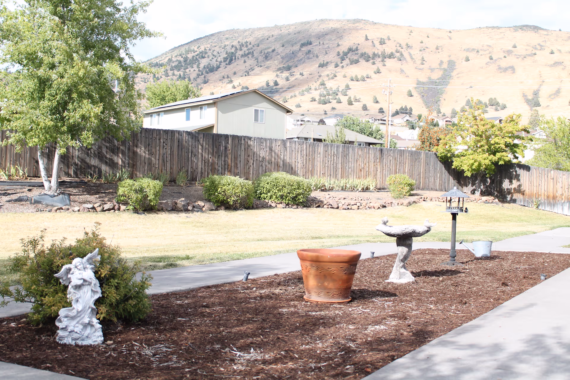 Outdoor garden area with a small statue, a large empty terracotta pot, a birdbath, and a bird feeder on a mulched bed next to a concrete walkway. There is a wooden fence and some bushes in the background, with a hill and houses visible beyond the fence under a partly cloudy sky.