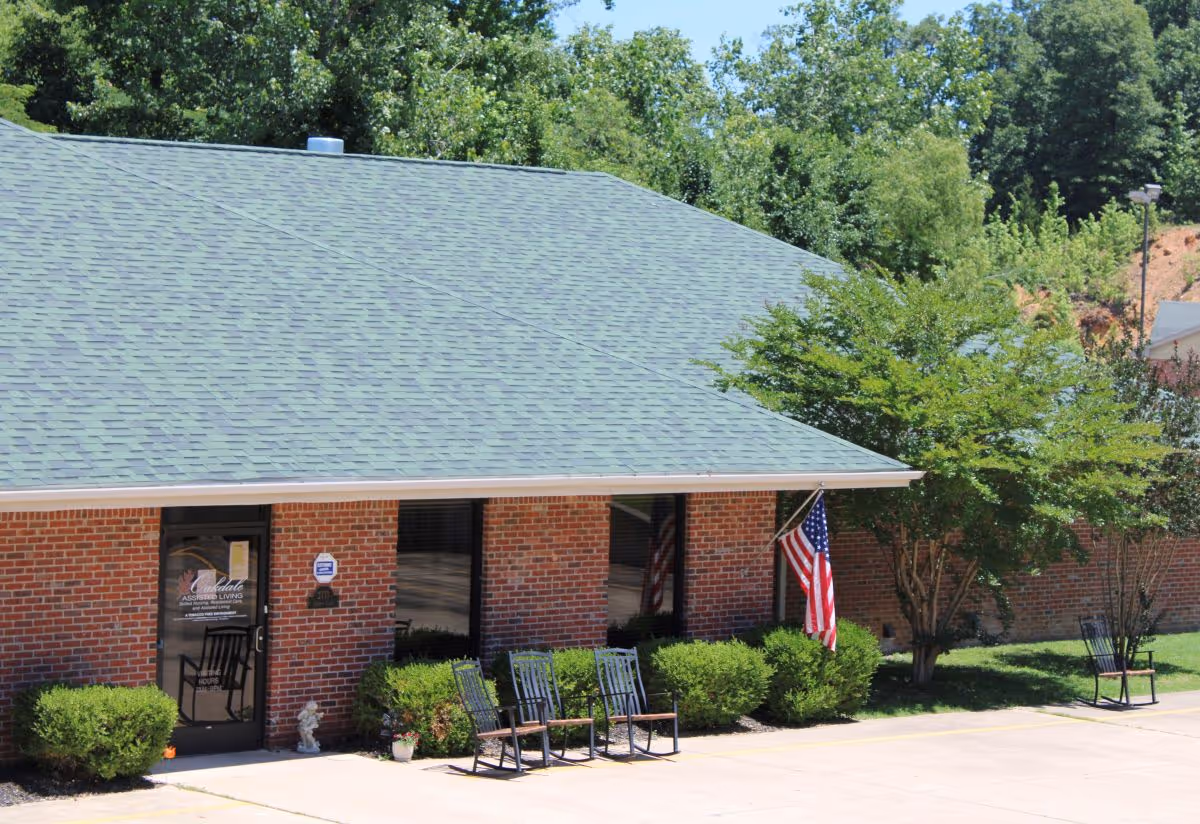 Exterior view of Oakdale Care Center showing a brick building with a green shingled roof, several windows, an American flag, bushes, and four black rocking chairs on the sidewalk in front of the entrance.