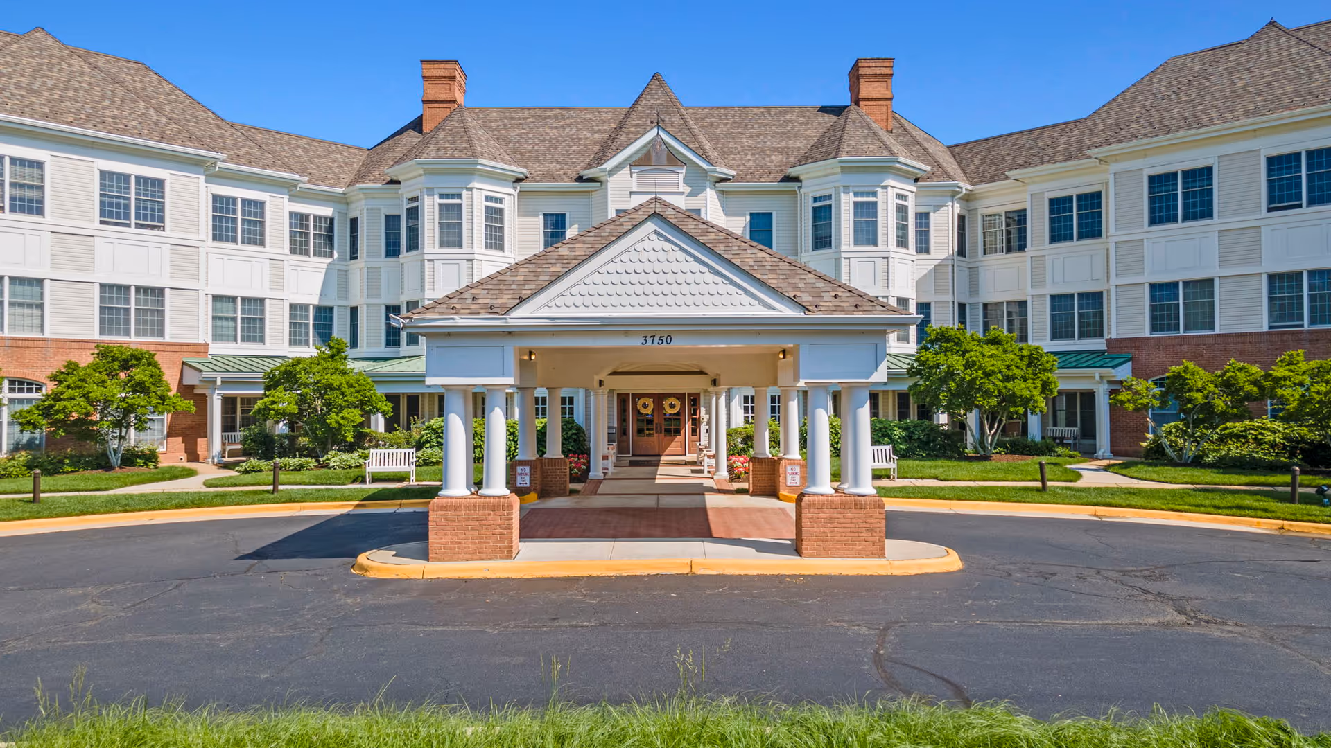 Front exterior view of a large senior living facility building with white siding and brick accents, multiple windows, and a covered entrance supported by white columns. The building is surrounded by green shrubs and trees under a clear blue sky.