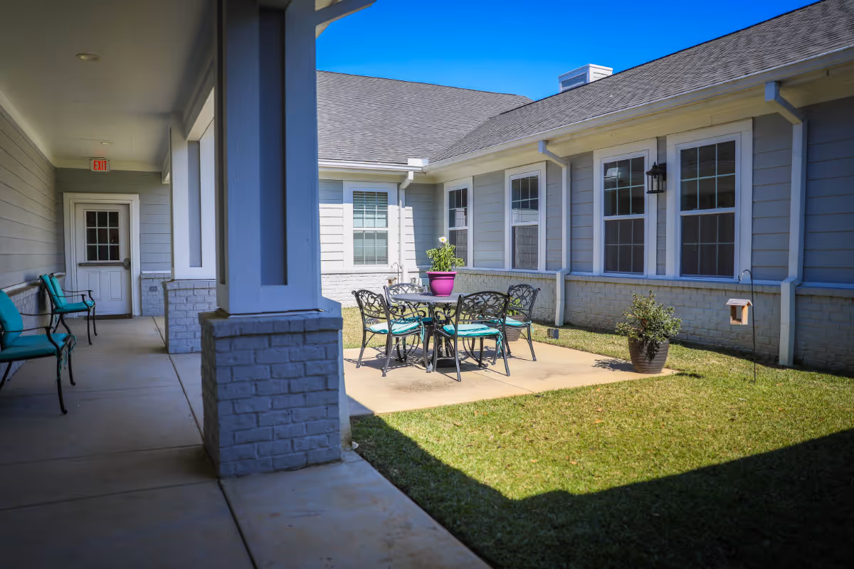 Outdoor courtyard area of a senior living facility with a patio table and four chairs with teal cushions. The courtyard is surrounded by a building with multiple windows and a covered walkway with additional seating along the wall. There is a potted plant on the table and another on the grass nearby under a clear blue sky.