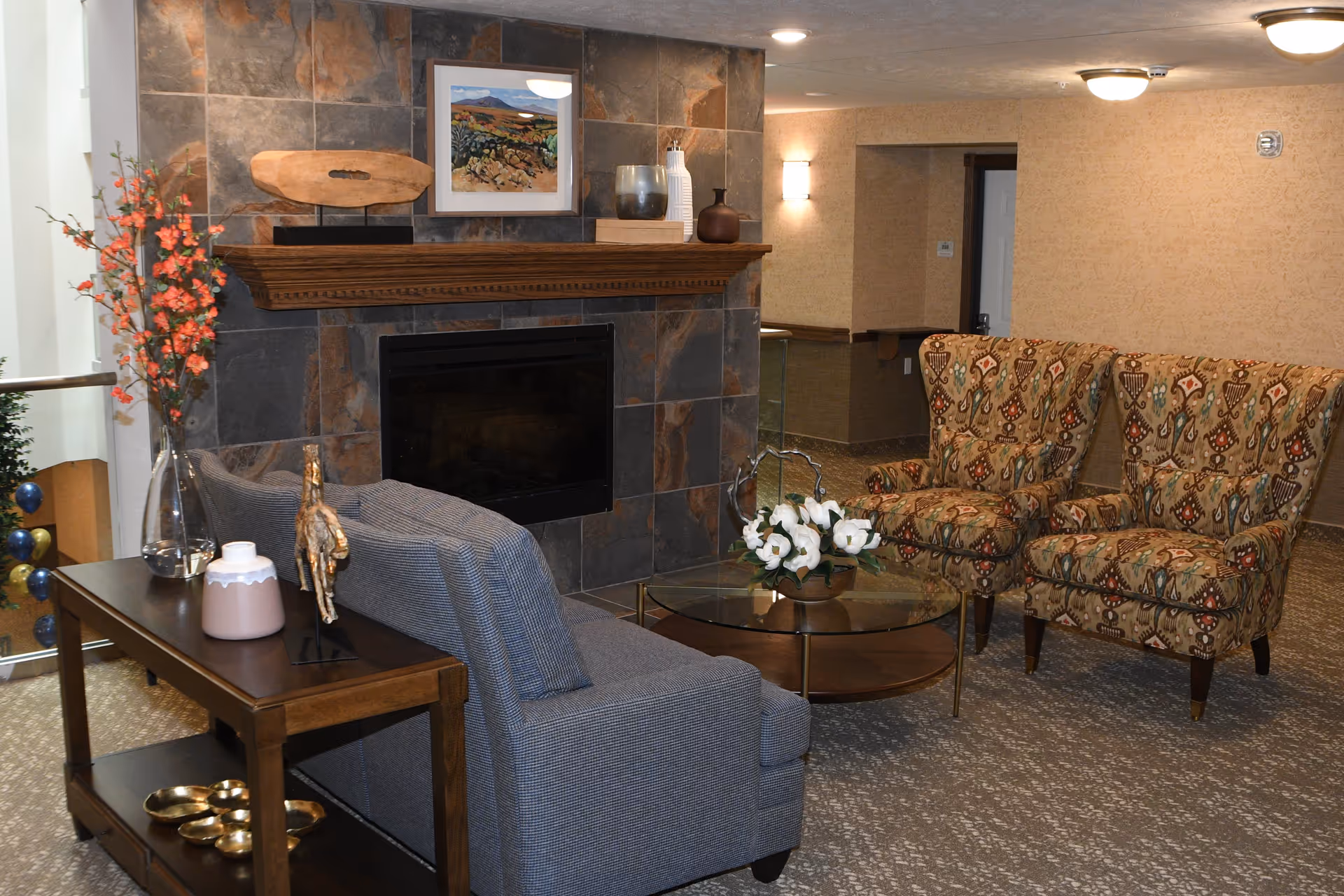 A cozy sitting area in a retirement community featuring a stone fireplace with a wooden mantel decorated with art and vases. In front of the fireplace is a gray upholstered sofa, a wooden side table with decorative items, a glass coffee table with a flower arrangement, and two patterned armchairs. The room has warm lighting and carpeted flooring.
