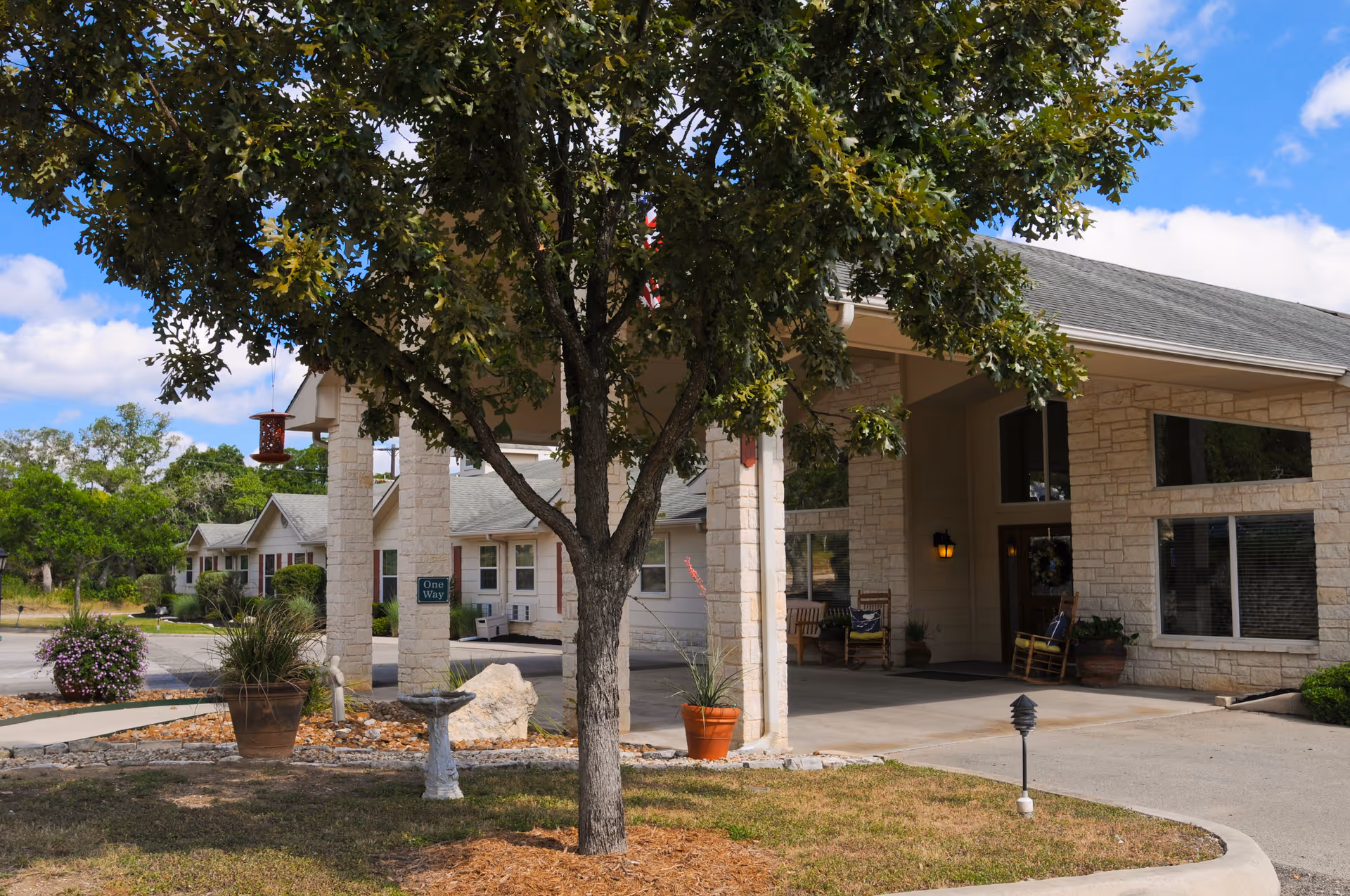 Exterior view of a senior living facility entrance with a covered driveway, stone pillars, a tree in the foreground, potted plants, and a birdbath. The building has large windows and a welcoming porch area with chairs.