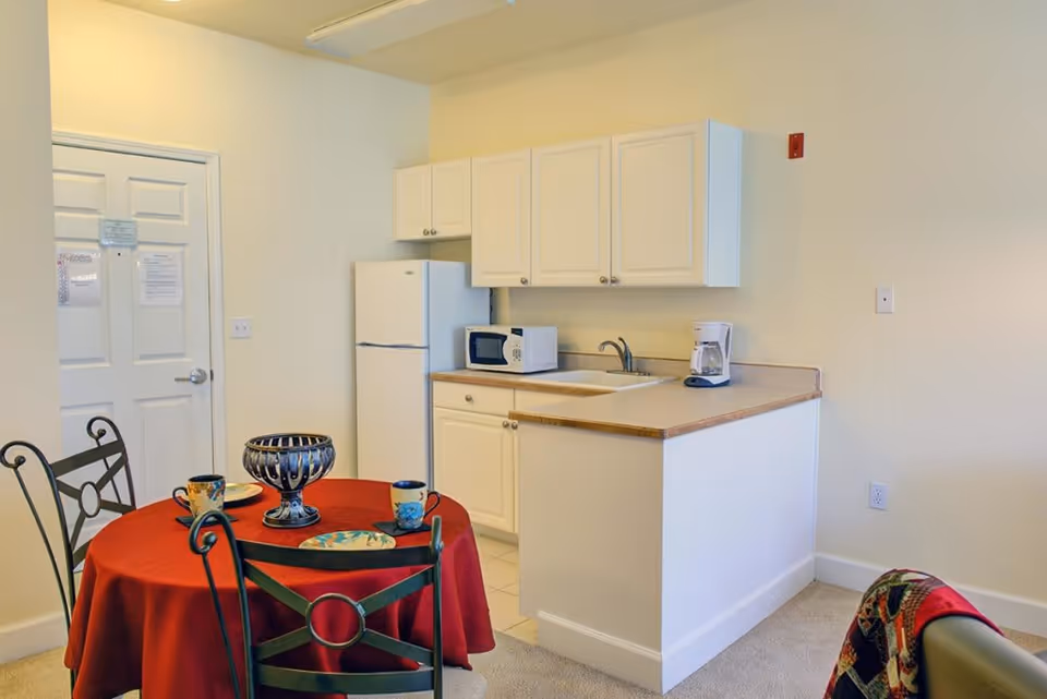 Small kitchen area with white cabinets, a refrigerator, microwave, coffee maker, and a sink. In front of the kitchen is a round table covered with a red tablecloth, set with plates and mugs, and surrounded by black metal chairs. A door is visible in the background.