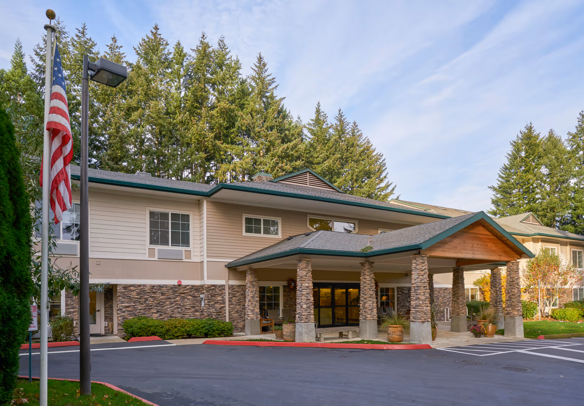 Exterior view of Woodland Heights Assisted Living facility showing the main entrance with a covered driveway supported by stone pillars, surrounded by trees and landscaping, with an American flag on a pole to the left.