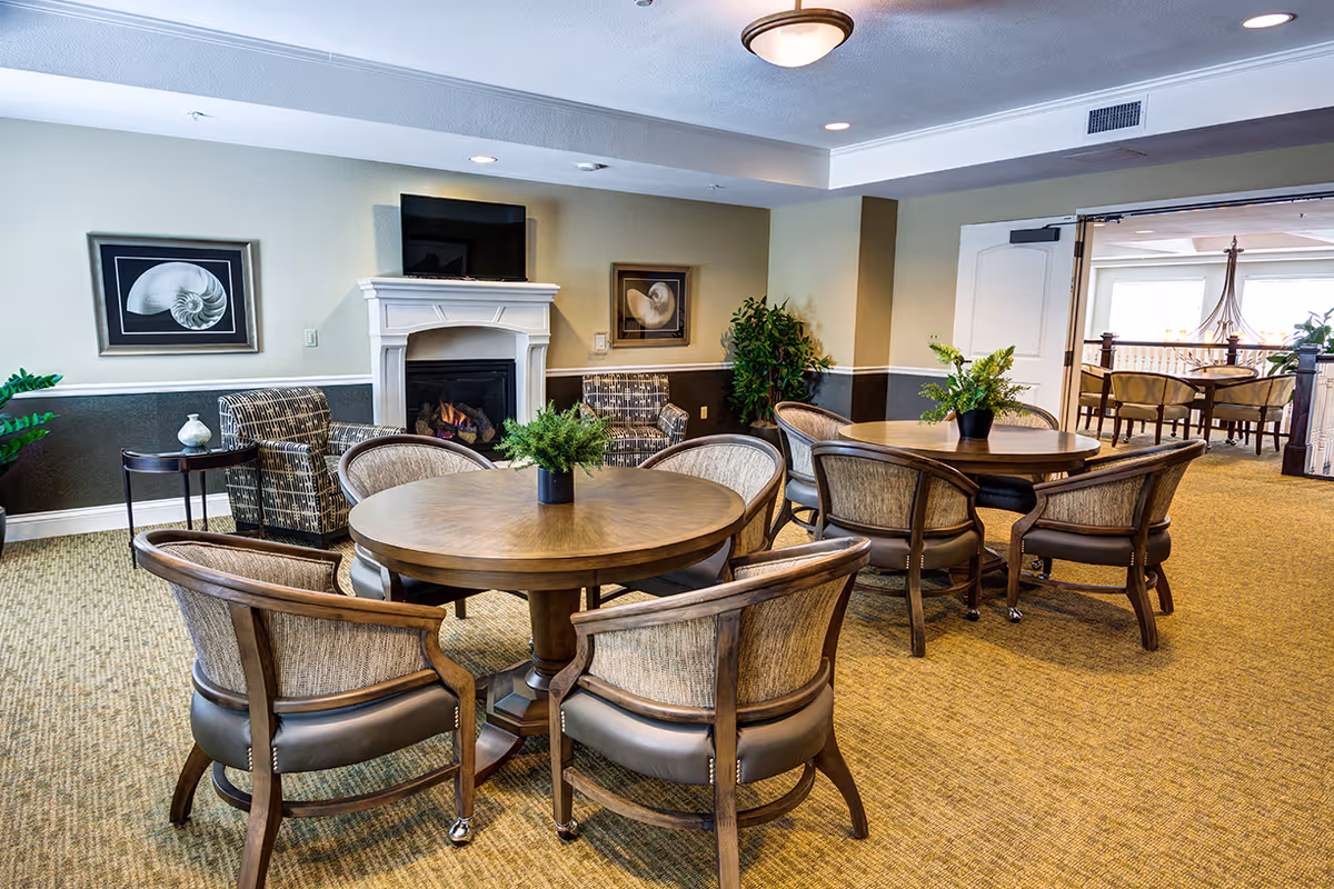 A cozy senior living common area with round wooden tables surrounded by cushioned chairs. There is a fireplace with a mounted TV above it, two framed shell pictures on the wall, and two patterned armchairs beside the fireplace. The room has beige walls with a darker lower half, carpeted floor, and potted plants for decoration. An open doorway leads to another room with more tables and chairs.