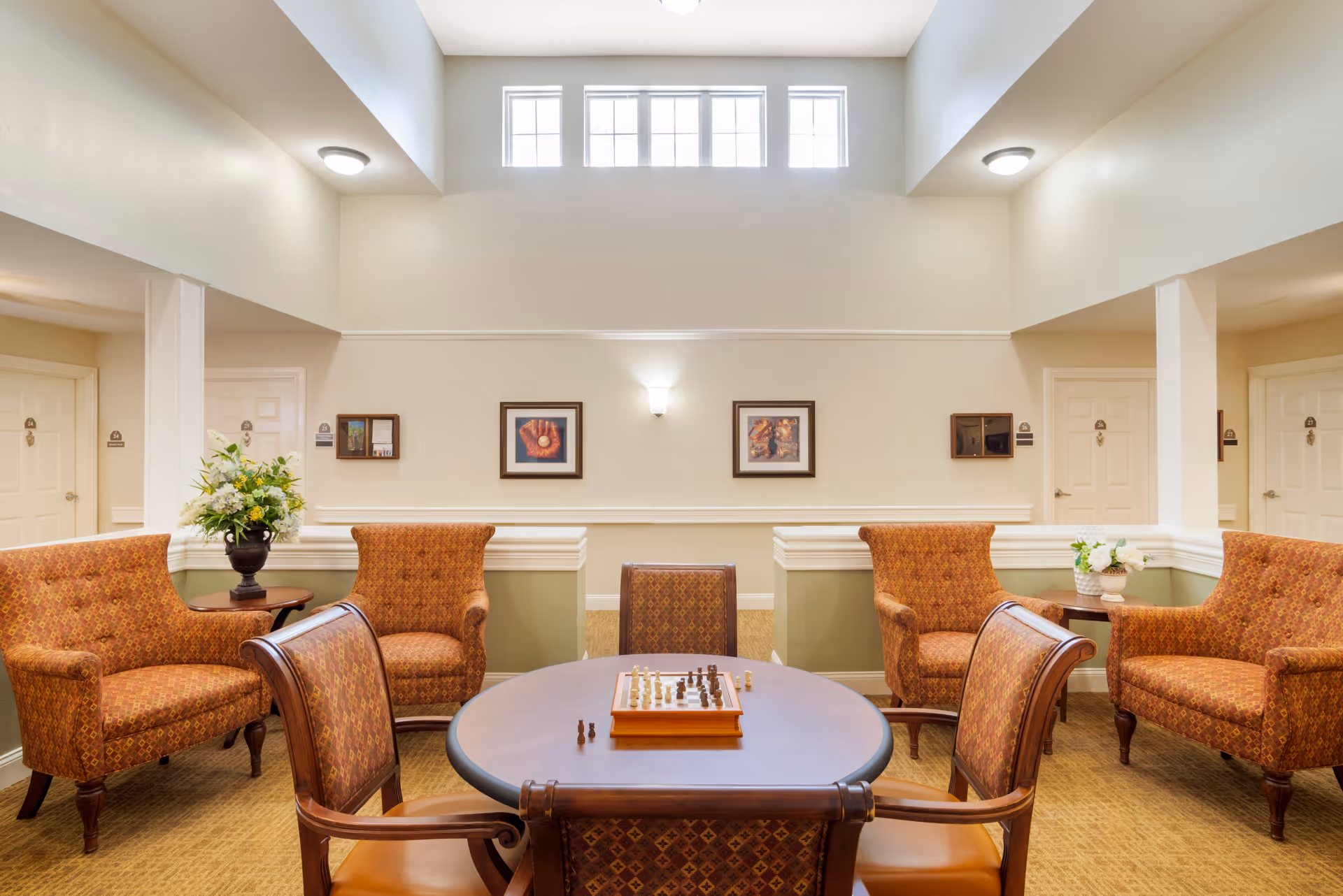 A well-lit common area with a high ceiling and upper windows. The room features a round table with a chessboard and chess pieces on it, surrounded by four upholstered chairs with patterned fabric. Around the perimeter are four matching armchairs and two small tables with flower arrangements. The walls are decorated with framed artwork and there are several doors visible in the background.