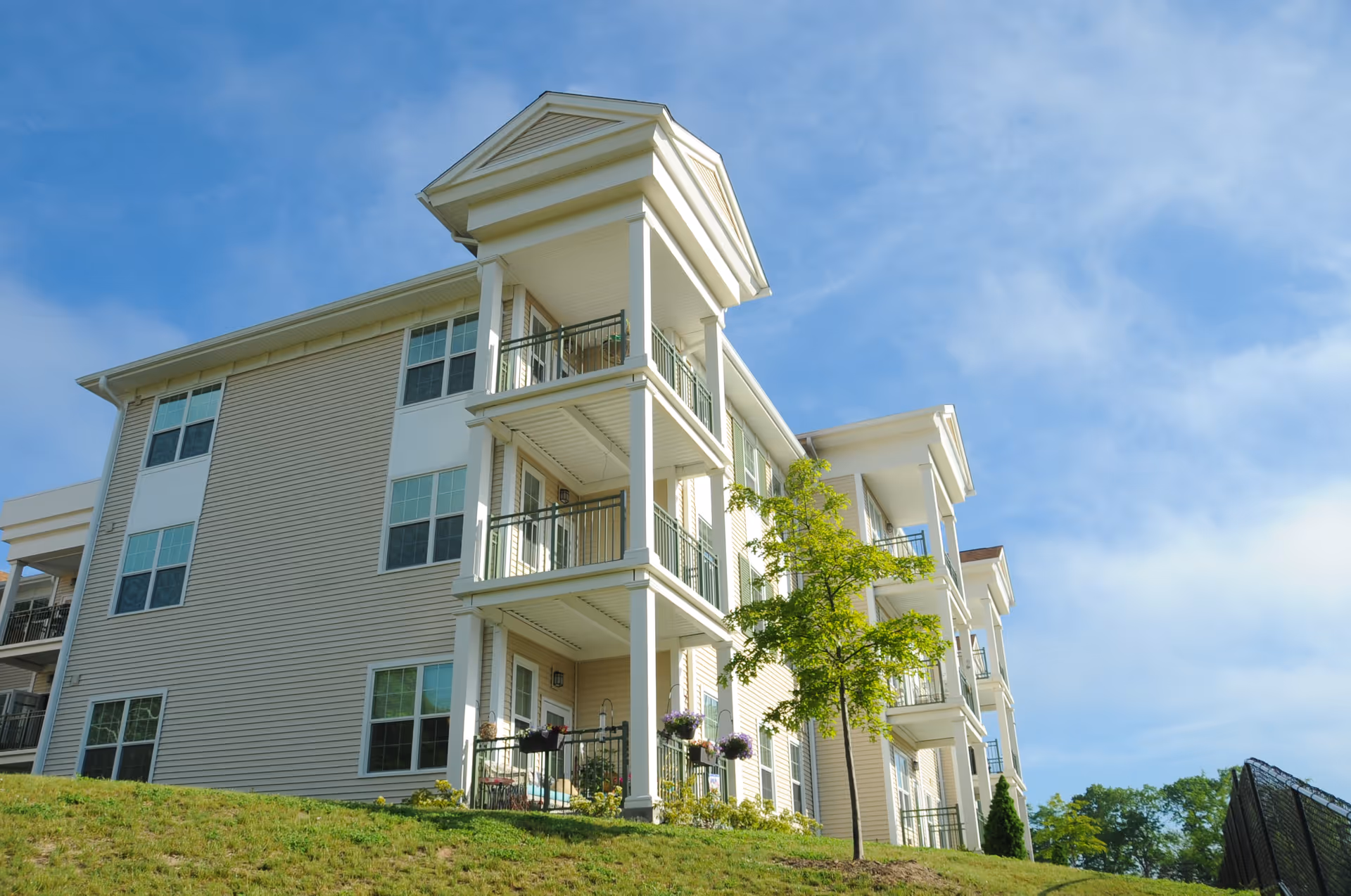Exterior view of a beige multi-story senior living building with balconies and a grassy lawn under a blue sky.