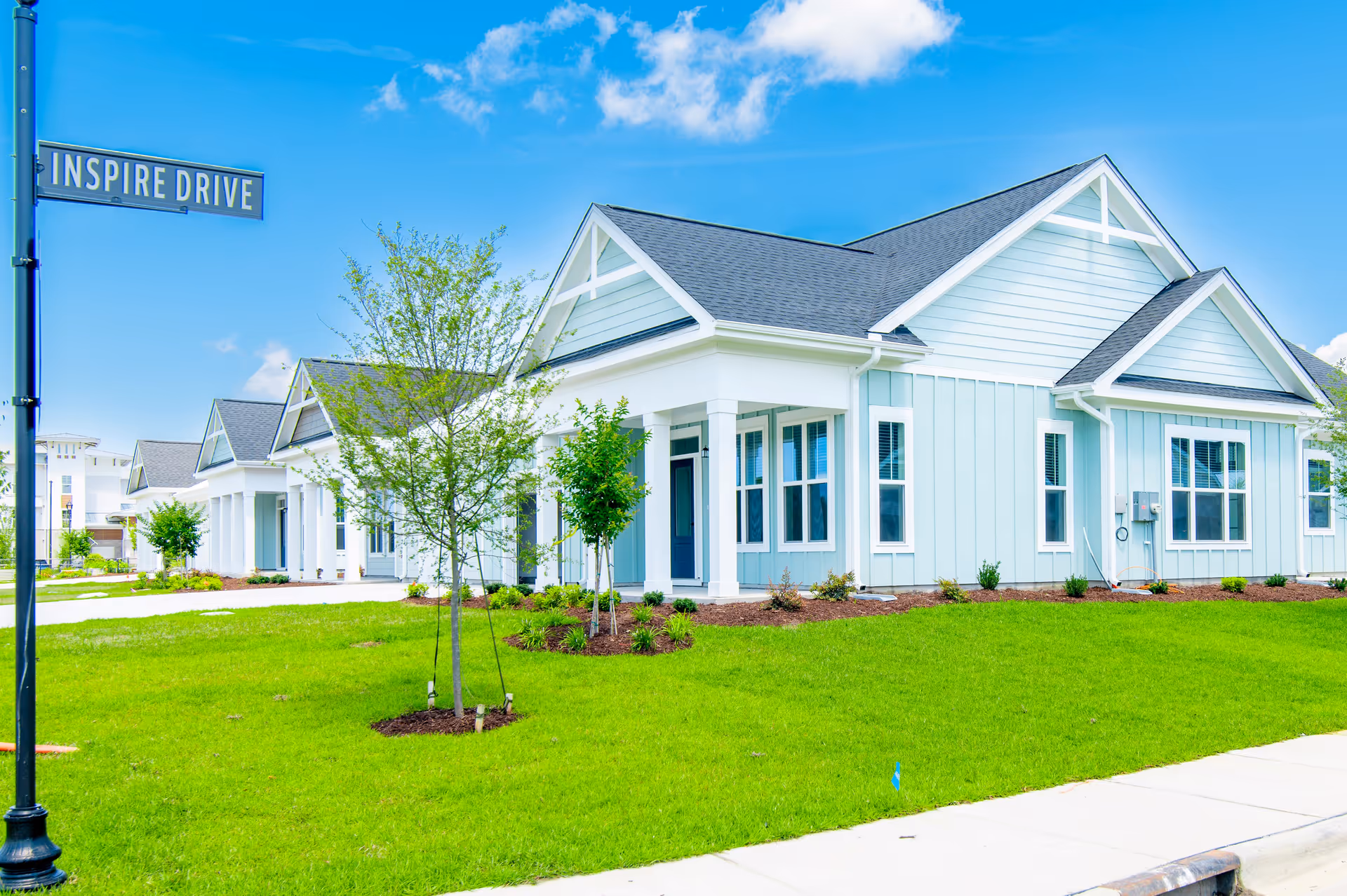 Exterior view of a light blue and white residential building with a dark gray roof, surrounded by green grass and small trees under a bright blue sky with some clouds. A street sign reading 'INSPIRE DRIVE' is visible on a black lamp post in the foreground.