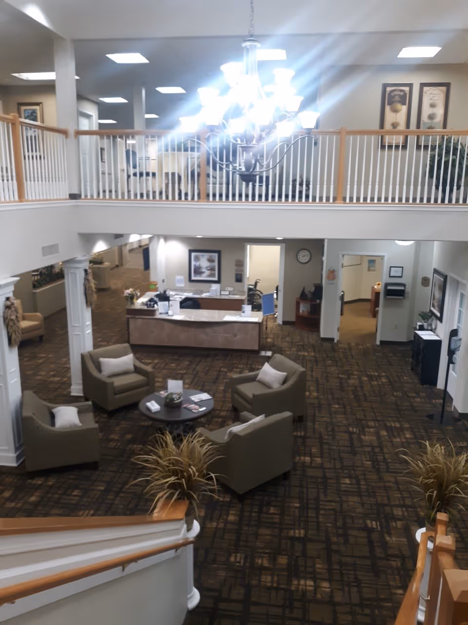 Interior view of a senior living facility lobby area with a seating arrangement of four armchairs around a round table. The space features a reception desk in the background, a large chandelier hanging from the ceiling, and a second-floor balcony with white railings. The carpet has a patterned design, and there are plants and framed artwork on the walls.