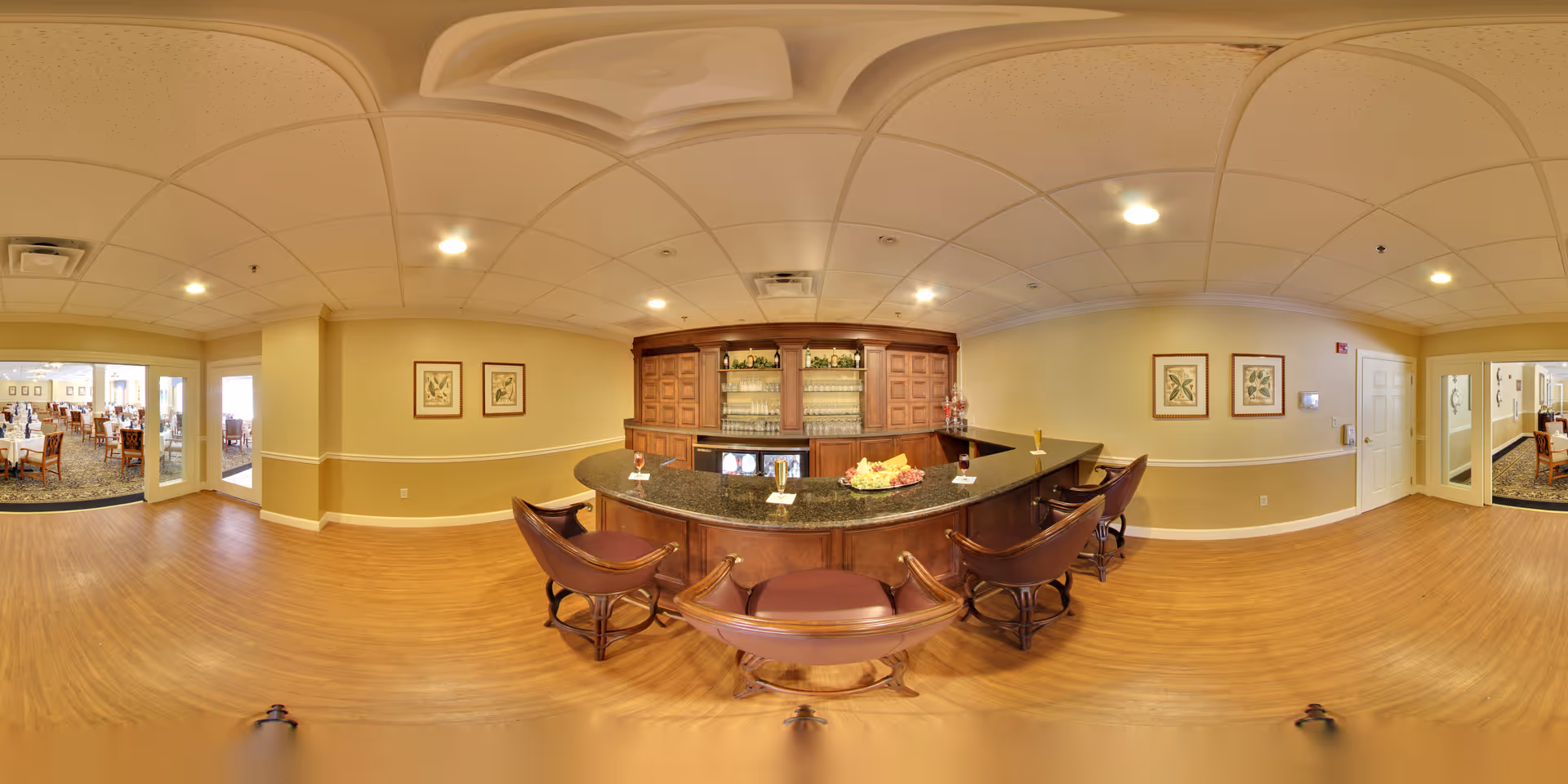 A spacious room with a wooden bar counter and four bar stools with leather seats. Behind the bar is a wooden cabinet with glass shelves holding bottles and glassware. The room has light yellow walls with framed botanical prints and a wood floor. To the left and right, there are doorways leading to a dining area with tables and chairs.