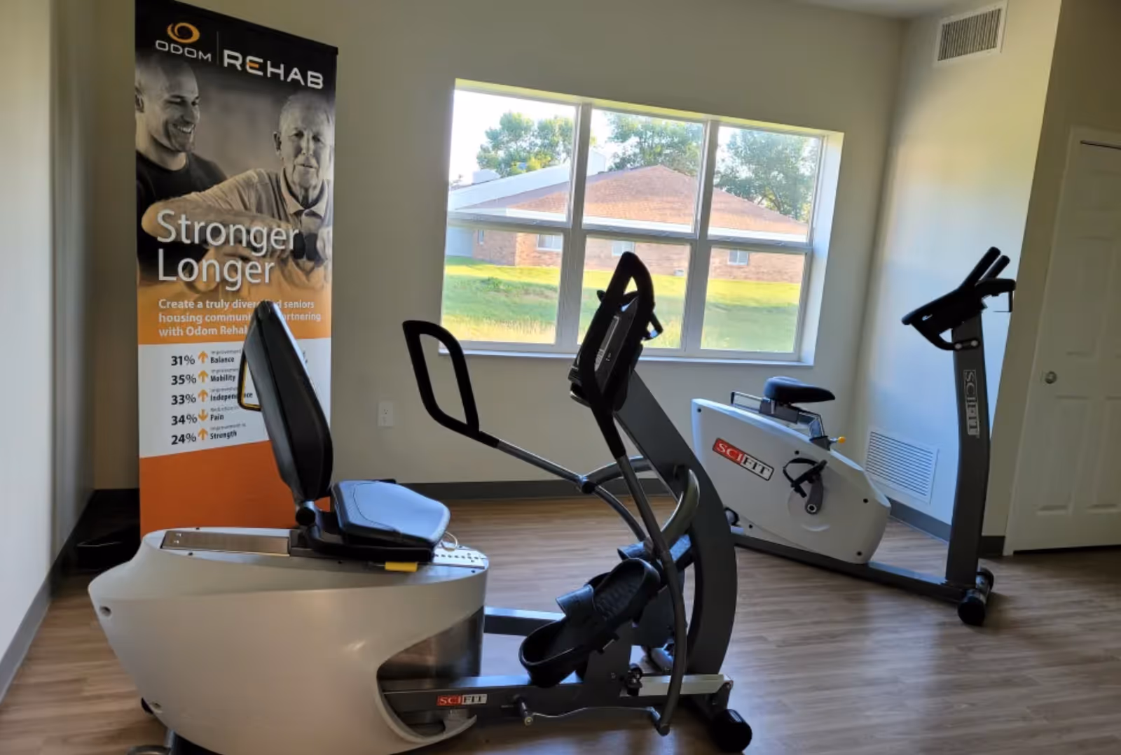 A small exercise room with two SciFit stationary exercise bikes positioned on a wooden floor. A large window lets in natural light and shows a view of a grassy area and a brick building outside. To the left, there is a vertical banner with the heading 'Odom Rehab' and the slogan 'Stronger Longer' along with some statistics about improvements in balance, mobility, independence, pain, and strength.