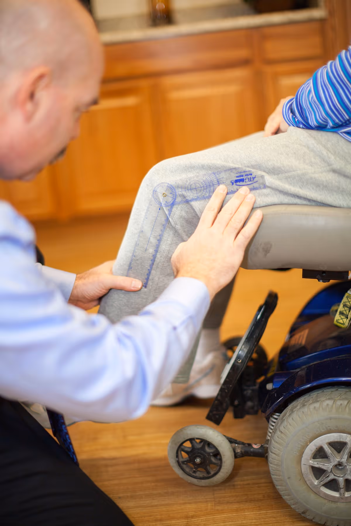 A man in a light blue shirt is examining or measuring the knee of a person sitting in a wheelchair, who is wearing gray sweatpants and white socks. The setting appears to be indoors with wooden flooring and wooden cabinets in the background.