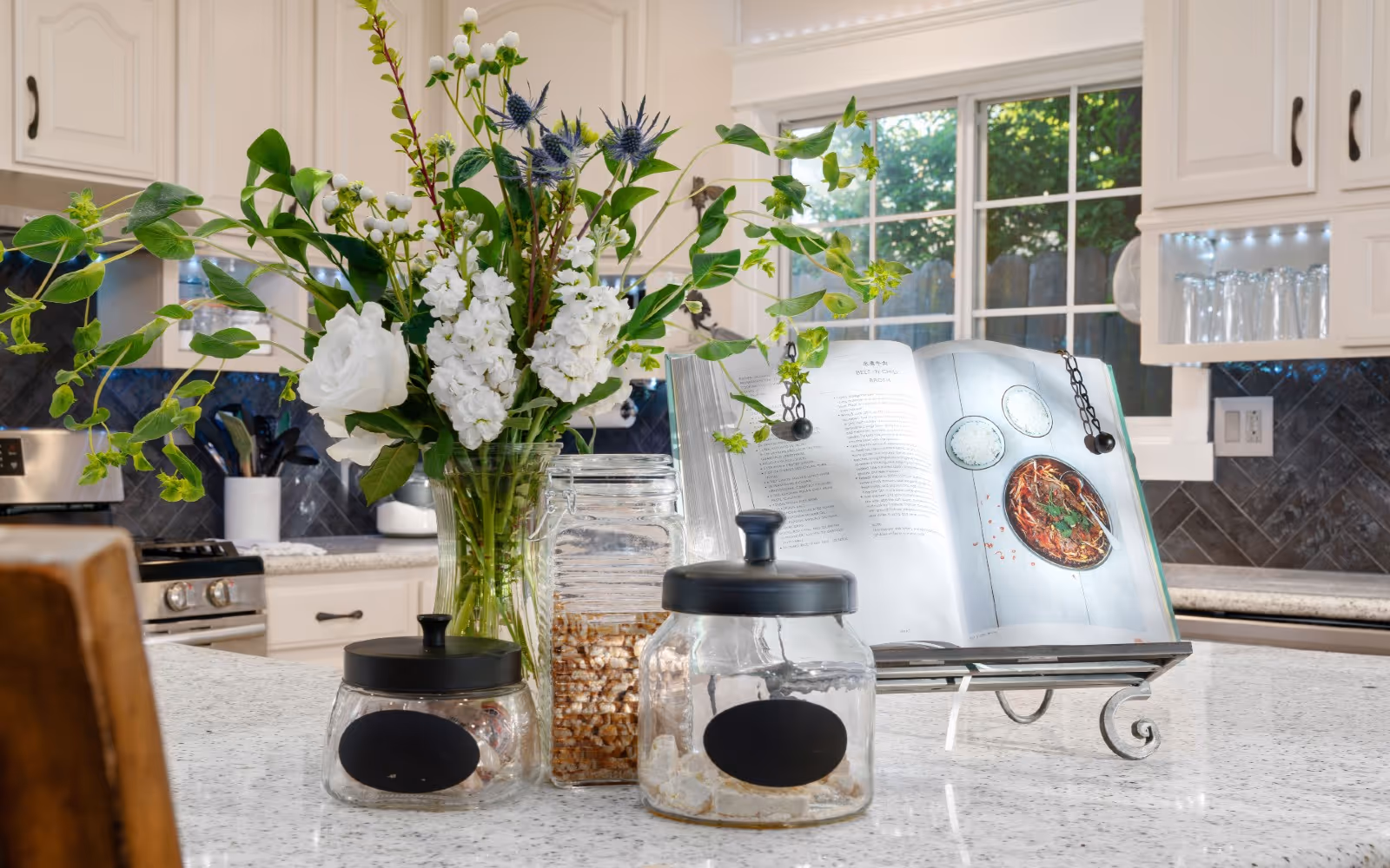 Kitchen island with a vase of white flowers, glass jars, and an open cookbook on a stand in a bright white kitchen.