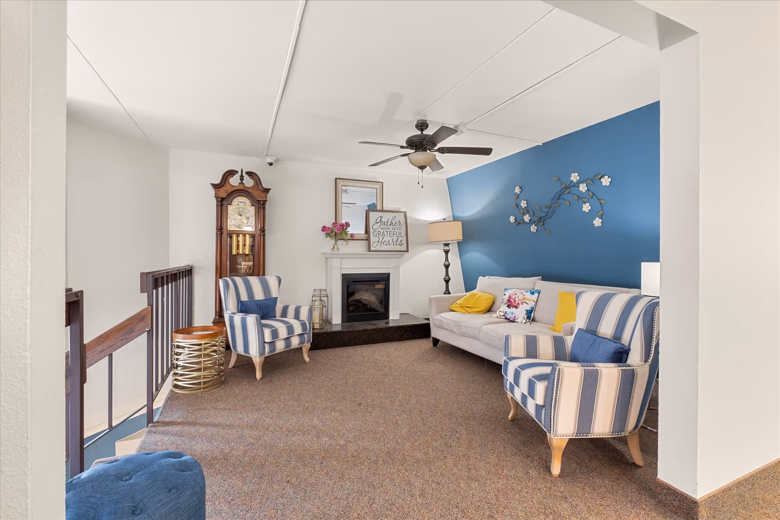 A cozy living room area with a beige sofa adorned with colorful pillows, two blue and white striped armchairs, a tall wooden grandfather clock, a white fireplace with decorative items on the mantel, a floor lamp, and a ceiling fan. The back wall is painted blue and decorated with a floral metal wall art.