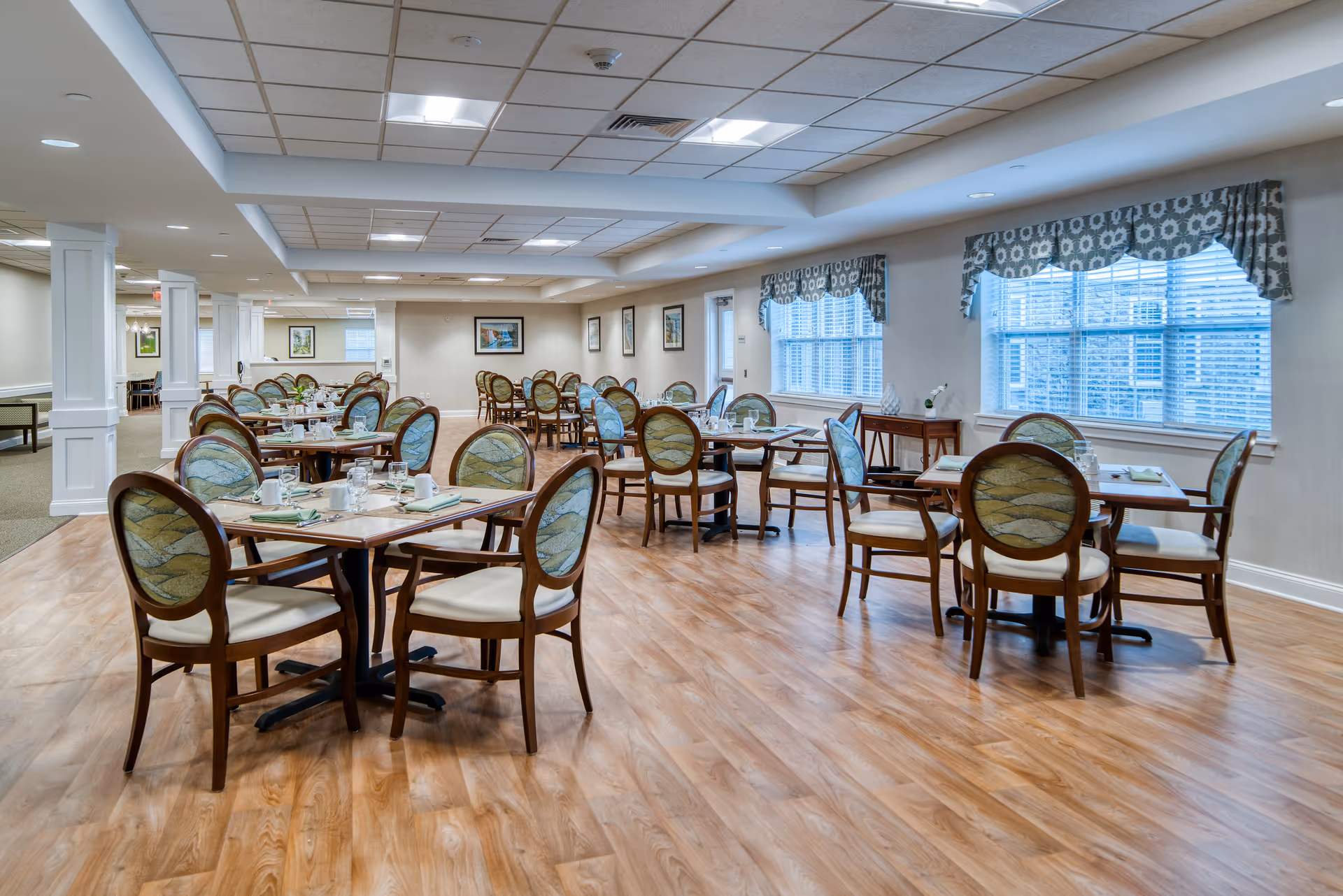 A spacious dining room in a senior living facility with multiple wooden tables and chairs arranged neatly. Each table is set with cups, glasses, napkins, and utensils. The room has large windows with patterned valances allowing natural light to enter, light-colored walls, framed artwork, and a wood-patterned floor.