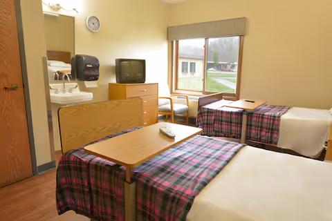A nursing facility room with two single beds covered in plaid blankets, a wooden bedside table with a remote control, a small TV on a dresser, two chairs by a window with a view outside, and a sink with a mirror and soap dispenser on the wall.