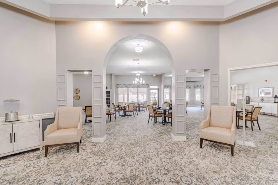 Bright and spacious common area in a senior living facility with patterned carpet, two beige armchairs on either side of a white cabinet with a water dispenser, and an arched entryway leading to a dining area with multiple tables and chairs. Large windows in the background allow natural light to fill the room.