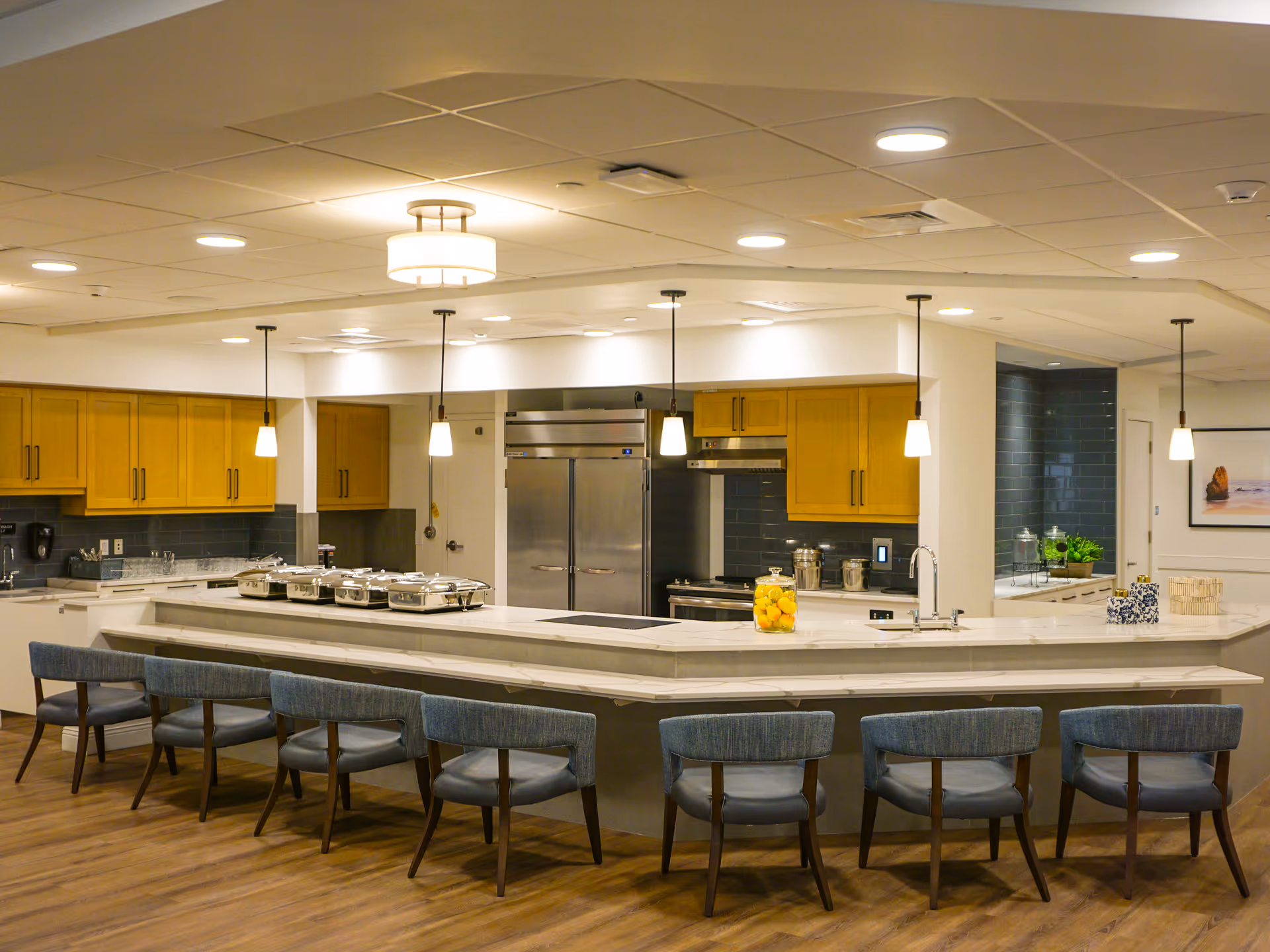 Bright communal kitchen with a long marble serving counter, barstools, pendant lights, stainless steel appliances and wooden cabinets.