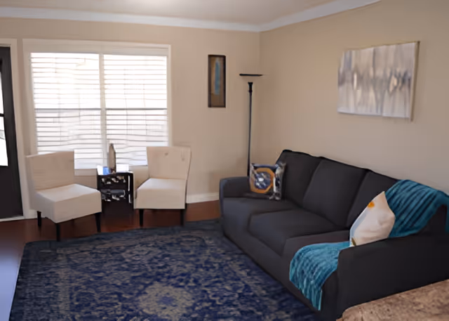 A cozy living room with a dark gray sofa adorned with two decorative pillows and a teal throw blanket. Two beige armchairs are positioned near a window with white blinds, separated by a small dark wooden side table. The room features a tall floor lamp, a framed artwork on the wall above the sofa, and a large blue patterned rug covering the wooden floor.