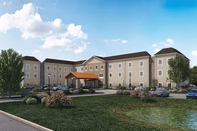 Exterior view of a three-story senior living facility building with beige walls and a stone accent around the entrance. The entrance has a covered porch with a brown roof. There are several cars parked in front, landscaped bushes, trees, and a small pond in the foreground under a partly cloudy sky.