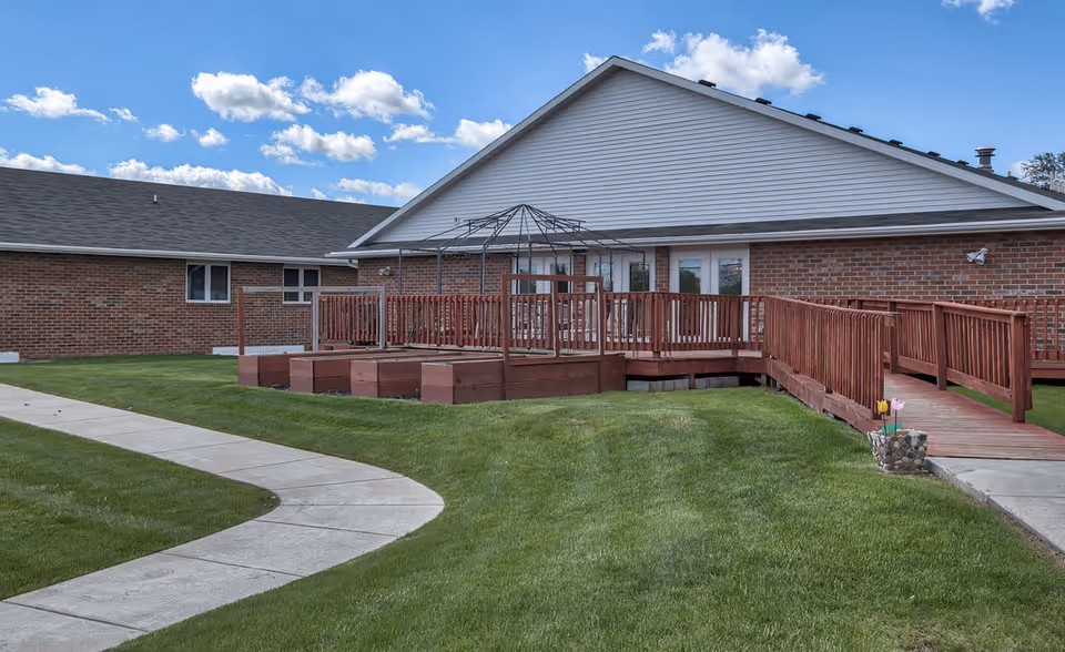 Outdoor view of a single-story brick building with a wooden deck and ramp surrounded by green grass and a curved concrete walkway under a blue sky with scattered clouds.