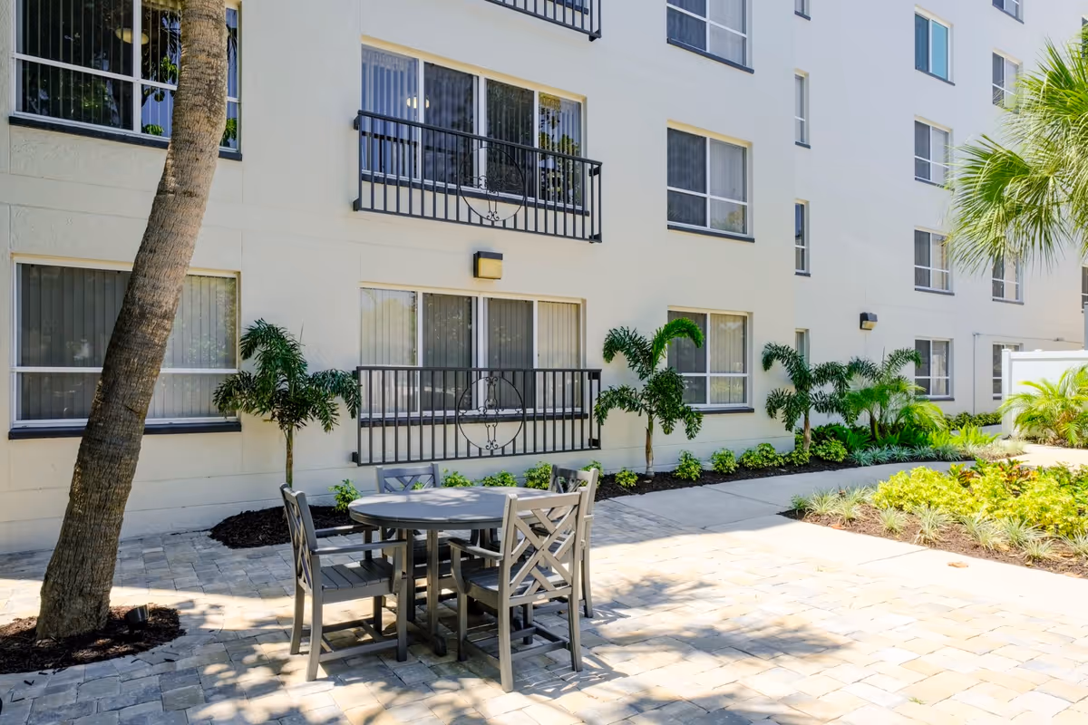 Outdoor patio area at Grand Villa of Sarasota with a round table and four chairs on a paved surface, surrounded by palm trees and plants, adjacent to a multi-story building with several windows and small balconies.